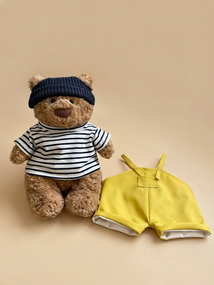Bartholomew Bear plush in a striped shirt beside yellow overalls and a navy hat